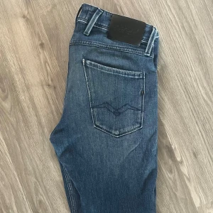 Replay blå jeans slim fit herr - Snygga blå jeans från Replay i slim fit-modell. Jeansen har klassisk femficksdesign, svarta läderpatch bak och är tillverkade i hållbar bomull. Snygga slitningar och detaljer på bakfickorna ger en modern look.