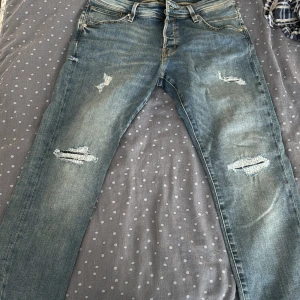 Slim Glenn jeans från Jack & Jones - Säljer ett par blå Slim Glenn jeans från Jack & Jones med slitna detaljer och snygga slitningar på benen. Jeansen har klassisk femficksdesign, normal midja och smal passform. Perfekta för dig som gillar en avslappnad men trendig look. 