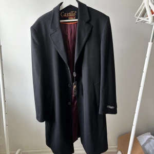 Svart rock från Castillo Italy i kashmir - Stilren svart rock från Castillo Italy med klassisk krage och knäppning framtill. Insidan har ett vinrött, randigt foder som ger en snygg kontrast. Jackan är tillverkad i ull och har två fickor på sidorna. Perfekt för dig som gillar en clean och tidlös look. Priset kan diskuteras vid snabb affär 