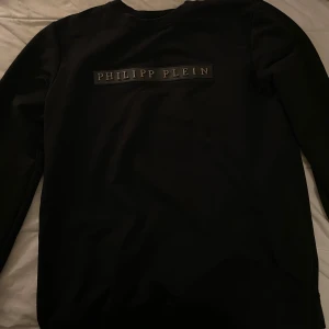 Svart Philipp Plein sweatshirt - Svart sweatshirt från Philipp Plein med logga broderad på bröstet och diskret PP-detalj på ärmen. Tröjan har rund halsringning och är tillverkad i mjuk bomull med lite stretch för extra komfort. Perfekt för dig som gillar stilrena och exklusiva plagg.