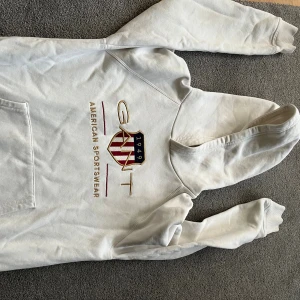 Vit hoodie från GANT med broderad logga - Vit hoodie från GANT med broderad logga och sköld på bröstet. Klassisk känguruficka framtill och huva. Mjuk insida och ribbade muddar vid ärmslut och nederkant. Perfekt för en chill och sportig look.