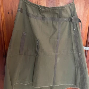 Olivgrön utilitykjol med dragkedja - Snygg olivgrön utilitykjol med rå kant, dekorativa band och metallöljetter i midjan. Kjolen har en praktisk ficka med dragkedja och bälte i midjan för extra edge. Perfekt för dig som gillar en cool och avslappnad stil. Står 42 men skulle säga att det passar 38-40