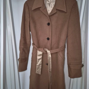 Beige kappa från Tiger of Sweden - Elegant beige kappa från Tiger of Sweden med klassisk krage, knappar framtill och ett bälte i midjan. Jackan har långa ärmar med knappdetaljer och är fodrad med ett glansigt tyg. Perfekt för dig som gillar stilrena och tidlösa plagg.