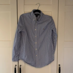 Blåvit randig skjorta från Ralph Lauren - Snygg skjorta från Ralph Lauren i blått och vitt med klassiskt randigt mönster. Skjortan har knappar hela vägen, button-down krage och den ikoniska broderade loggan på bröstet. Tillverkad i mjuk bomull och har lång ärm. Perfekt för dig som gillar stilren och tidlös design. Nypris 2000kr 