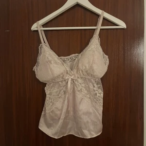 Ljusrosa spetsig linnetopp med rosett - Säljer en ljusrosa linnetopp med tunna axelband och vacker spetsdetalj över bysten och sidorna. Toppen har en liten rosett framtill och är tillverkad i glansigt satinliknande material med subtila ränder. Perfekt för dig som gillar romantisk och feminin stil.