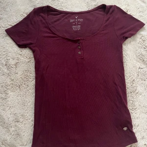 Vinröd ribbad topp från American Eagle - Superskön vinröd ribbad topp från American Eagle med rundad halsringning och tre knappar framtill. Kortärmad och figurnära passform, perfekt för dig som gillar enkel men snygg stil. Soft & Sexy-kollektionen, stretchigt bomullsmaterial.