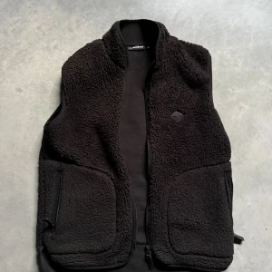 Svart fleeceväst från J.Lindeberg - Mysig svart fleeceväst från J.Lindeberg med hög krage och två stora fickor med dragkedja framtill. Västen har en enkel, stilren design och är perfekt att slänga på sig när det är kyligt ute. Materialet är mjukt och fluffigt, vilket gör den extra skön.