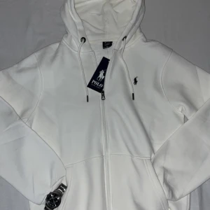 Vit Ralph Lauren full zip hoodie  - Säljer en sprillans ny clean vit hoodie från Polo Ralph Lauren med dragkedja och huva. Tröjan har snörning i huvan, två fickor fram och klassisk logga på bröstet. Perfekt för dig som gillar stilren och enkel streetwear.