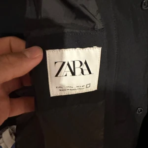 Svart quiltad bomberjacka från Zara - Svart bomberjacka från Zara med quiltad framsida och ribbad krage. Jackan har en stilren look med tryckknappar och raka linjer. Perfekt för dig som gillar minimalistisk stil och vill ha en jacka som funkar till många outfits.