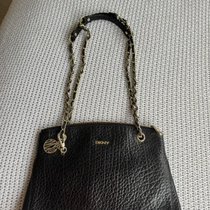 Svart axelväska från DKNY i skinn - Snygg svart axelväska från DKNY i äkta skinn med grov textur. Väskan har guldfärgade detaljer, kedja med läderflätning och ett DKNY-hänge. Stilren och klassisk form, perfekt för dig som gillar en chic look.