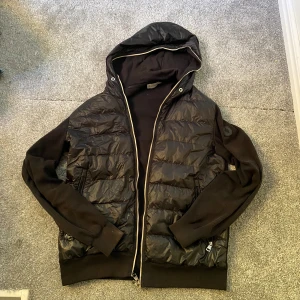 Moncler cardigan  - Svart pufferjacka från Moncler med huva och dragkedja framtill. Jackan har quiltad framsida i glansigt material. Priset är ej hugget i sten. Fler bilder går att få. XL juniorstorlek så den sitter som en M eller L
