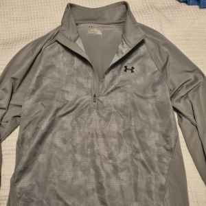 Grå half zip tröja från Under Armour - Grå långärmad tröja från Under Armour med diskret mönster och svart logga på bröstet. Söm har gått upp lite Under ena ärmen, därav priset. 