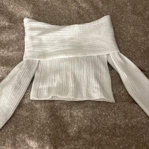 Vit ribbad offshoulder topp NA-KD - Superfin vit ribbad topp från NA-KD med offshoulder-design och långa ärmar. Materialet är mjukt och stretchigt, vilket gör den bekväm att bära. Den har en croppad passform och bred kant upptill som ger en snygg siluett.