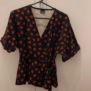 Blommig omlottblus från Gina Tricot - Säljer en svart omlottblus från Gina Tricot med röda och rosa blommor. Blusen har korta ärmar, v-ringning och knyt i midjan. Perfekt för dig som gillar färgglada mönster och en feminin look.