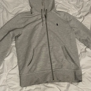 Grå hoodie från Polo Ralph Lauren - Snygg ljusgrå hoodie från Polo Ralph Lauren med dragkedja framtill, huva med snörning och klassisk logga på bröstet. Tröjan har två fickor och är tillverkad i mjuk bomullsmix. Perfekt för chill dagar eller när du vill ha en clean look.