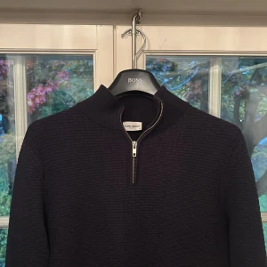 Svart stickad half zip-tröja John Henric - Mörk, stickad tröja från John Henric med snygg struktur och half zip-dragkedja vid halsen. Tröjan har hög krage och är perfekt för kyliga dagar. Klassisk och stilren modell som passar till det mesta.