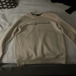 Beige Sail Racing sweatshirt M - Snygg beige sweatshirt från Sail Racing i storlek M. Tröjan har rund hals, lång ärm och svart logga framtill. Tillverkad i mjuk bomull och polyester för en skön känsla. Perfekt för dig som gillar stilren och enkel design.
