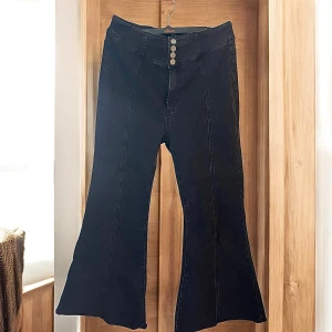 svarta bootcut jeans från Cider med hög midja storlek L - Snygga svarta bootcut jeans från Cider med hög midja och fyra silverfärgade knappar framtill. Byxorna har markerade sömmar längs benen och är tillverkade i ett stretchigt jeansmaterial som sitter bekvämt.