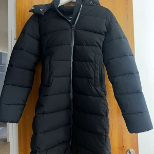 Svart lång pufferjacka från MNG XS - Lång svart pufferjacka från MNG med huva och dragkedja framtill. Jackan är quiltad och har en clean look med raka linjer. Materialet är vattenavvisande och andas, perfekt för kalla dagar. Snygg och funktionell för vintern. Knappast använd köpt för 1199kr. 