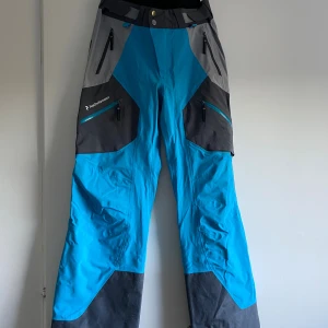 Peak Performance skalbyxor Gore-Tex - Blå och grå skalbyxor från Peak Performance med svarta detaljer. Byxorna har flera dragkedjeförsedda fickor, förstärkta benslut och är tillverkade i slitstarkt Gore-Tex-material. Perfekta för vinterns äventyr och outdoor-aktiviteter.
