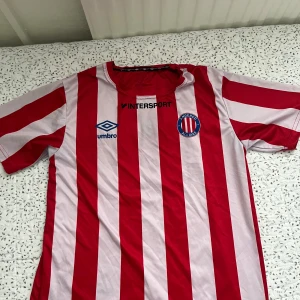 Rödvit randig Angered Allians fotbollströja - Säljer en klassisk fotbollströja . Materialet är lätt och andas, perfekt för match eller träning. Tryck med Intersport och Umbro-logga på bröstet.