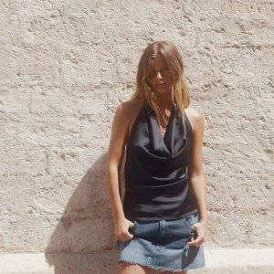 Svart halterneck satin topp - Snygg svart halterneck topp i satin med draperad v-ringning framtill och öppen rygg med knyt i nacken. Toppen har en elegant, glansig finish och sitter figurnära över midjan. Perfekt för dig som vill ha en chic och trendig look.