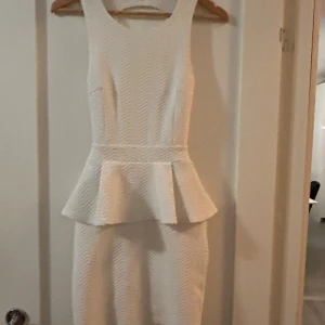 Vit peplumklänning med öppen rygg - Snygg vit kortklänning med peplumdetalj i midjan och cool öppen rygg med korsade band. Klänningen är ärmlös och har en strukturerad yta med diskret mönster. Perfekt för dig som vill sticka ut med en unik och trendig look. Funkar även till xs