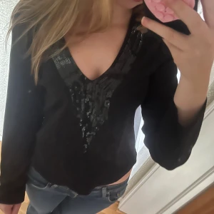 Svart långärmad topp med paljetter - Säljer en svart långärmad topp med djup v-ringning och snygga svarta paljettdetaljer längs ringningen och framtill. Toppen har en figurnära passform. Materialet känns mjukt. 