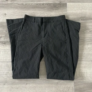 Svarta kostymbyxor med vita ränder - Snygga svarta kostymbyxor med smala vita ränder i klassiskt pinstripe-mönster, från Gina Byxorna har rak passform, bälteshällor och sidofickor. P