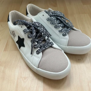 Vita sneakers med leopardmönstrade snören - Snygga vita sneakers med beige mockadetalj på tån, svarta glitterdetaljer på hälen och coola leopardmönstrade snören. Dekorerade med svarta och silvriga stjärnor på sidan. Platt sula och rund tå för skön passform.