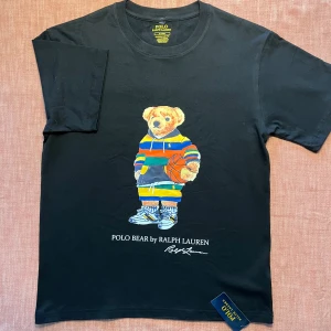 Svart Polo Bear t-shirt från Ralph Lauren - Svart t-shirt från Polo Ralph Lauren med ikoniska Polo Bear-trycket på bröstet. Björnen har färgglad hoodie och basketboll. T-shirten har rund hals, korta ärmar och är i slim fit. Materialet är mjuk bomull och märkeslappen sitter kvar.