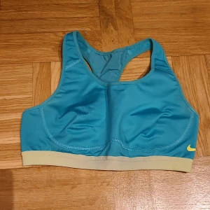 Turkos sport-bh från Nike Dri-FIT - Nike Dri-FIT sport-bh i turkos med ljusgul resår och liten gul Swoosh-logga framtill. Modellen är ärmlös med brottarrygg och tillverkad i ett snabbtorkande syntetmaterial som passar perfekt för träning.