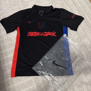 Svart FC Barcelona x Nike Cactus Jack tröja - Säljer en svart FC Barcelona x Nike Cactus Jack fotbollströja med röd och blåa detaljer på sidorna. Tröjan har korta ärmar, Dri-Fit material och coolt grafiskt tryck på bröstet. Nike-logga och FCB-emblem i svart med röd och blå outline.