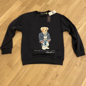 Svart sweatshirt Polo Bear Ralph Lauren - Svart sweatshirt från Ralph Lauren med ikoniskt Polo Bear-tryck framtill. Tröjan har rund hals, ribbade muddar och lång ärm. Materialet är mjuk bomull och passformen är normal. Perfekt för dig som gillar streetstyle och klassiska märken.