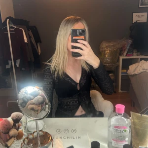 Svart spetstopp från NA-KD XS - Svart långärmad topp från NA-KD i spets med  mönster. Modellen är croppad med v-ringning och knappar framtill. Använd 1 gång! Köpt för 299kr.