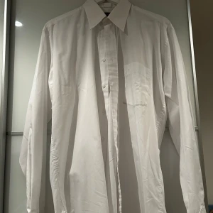 Vit skjorta från Melka Menswear *M* - Klassisk vit skjorta från Melka Menswear med lång ärm, knappar framtill och bröstficka. Skjortan har en stilren och tidlös design med normal passform och är tillverkad i mjuk bomull. Perfekt för dig som gillar clean och enkel stil.