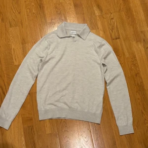 Ljusgrå merinoull pikétröja These Glory Days - Snygg ljusgrå pikétröja från These Glory Days i mjuk superior merinoull. Tröjan har klassisk krage, v-ringad hals och långa ärmar. Perfekt för dig som gillar stilrena och bekväma plagg med lite extra premiumkänsla. Modellen är 173cm lång och väger cirka 70kg.