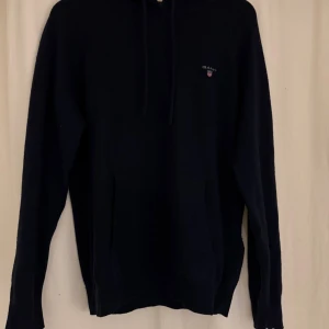 Mörkblå stickad kashmir hoodie från GANT - Mörkblå hoodie från GANT i en kashmir blend med huva och snörning. Klassisk logga på bröstet och praktisk magficka framtill. Långärmad och perfekt för kyliga dagar. Snygg och enkel design som funkar till jeans eller mjukis. Nypris 2600!