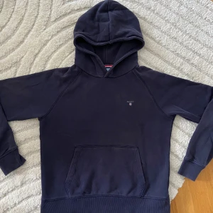 Mörkblå hoodie från GANT - Snygg mörkblå hoodie från GANT med klassisk huva och magficka. Tröjan har diskret logga på bröstet och ribbade muddar vid ärmslut och nederkant. Perfekt för chill eller häng med kompisar. Mjuk bomullskvalitet som känns skön mot huden. Köpt på Plick för 200:-- men passade ej här, så säljer vidare. Fint skick vad jag kan se. Hela muddar.