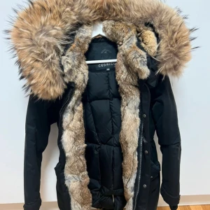 Svart dunjacka med päls från Cedrico - Snygg svart dunjacka från Cedrico med stor fluffig päls runt huvan och insida. Jackan har quiltad design, dragkedja och är perfekt för kalla vinterdagar. Pälsen är i naturliga bruna och beige toner som ger en lyxig känsla. Klassisk passform och långärmad. Helt ny utan prislapp. För liten mellan storlek 32/34