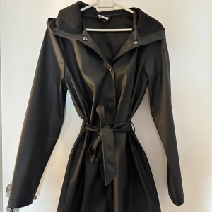 Svart regnjacka med huva och bälte - Svart regnjacka i syntetmaterial med huva, knappar framtill och knytskärp i midjan. Jackan har långa ärmar och en stilren, minimalistisk look. Perfekt för regniga dagar när du vill hålla stilen enkel och clean.