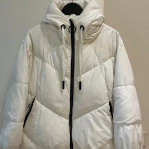  Ny Vit vinter jacka från Zara med huva - Vit pufferjacka från Zara med stor huva, svarta detaljer på dragkedja och snörning. Jackan har quiltad design, ribbade muddar och två fickor med dragkedja. Perfekt för kalla dagar och riktigt snygg till streetwear.