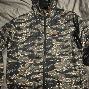 Camouflage vindjacka från Weekend Offender - Säljer en snygg vindjacka från Weekend Offender i camouflage-mönster med dragkedja framtill och huva. Jackan har flera praktiska fickor och coola detaljer som bruna snöröglor vid halsen. Perfekt för dig som vill sticka ut med streetstyle. Iconiskt och köpt i London