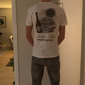 Vit Dolce Vita Portofino t-shirt Gildan - Snygg vit t-shirt från Gildan med Dolce Vita Portofino-tryck på ryggen. Motivet har en flaska, klocka, yacht och italiensk stad. Rund hals och korta ärmar, tillverkad i mjuk bomull. Perfekt för dig som gillar stilrena och unika prints.
