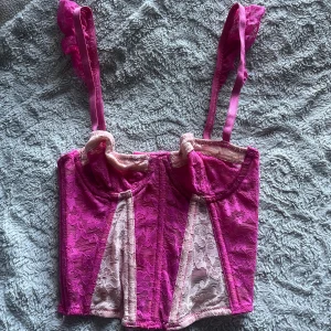 Rosa spetskorsett med axelband - Säljer en snygg rosa korsett från I AM GIA med justerbara axelband och hakknäppning bak. Korsetten har genomskinliga spetsdetaljer i rosa och ljusrosa, samt paneler som ger en cool kontrast. Perfekt för dig som vill sticka ut med en trendig look.