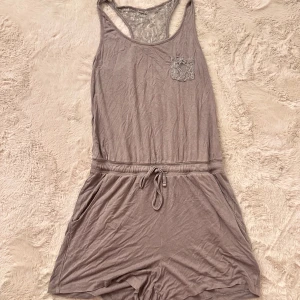 Grå jumpsuit med spetsdetaljer - Säljer en grå jumpsuit med shorts och snörning i midjan. Den har en snygg spetsdetalj både på ryggen och på bröstfickan. Materialet är mjukt och känns som bomull. Två fickor på sidorna och relaxed passform.