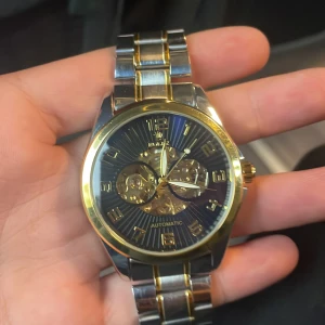 Automatisk klocka i guld och silver - Snygg automatisk klocka från Winner med urtavla i svart och detaljer i guld. Klockan har ett skelettur där du ser mekaniken, samt armband i silverfärgat stål med gulddetaljer. Perfekt accessoar för dig som gillar stilrena och lyxiga detaljer.