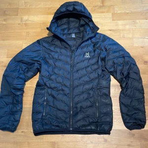 Svart dunjacka från Haglöfs - Svart dunjacka från Haglöfs i storlek S. Jackan har huva, två dragkedjeförsedda fickor och quiltad design. Perfekt för kyliga dagar och har en clean look med diskret logga på bröstet. Materialet är syntet och jackan är långärmad. Den har en liten defekt (se sista bilden) annars är den i ett bra skick. 8/10