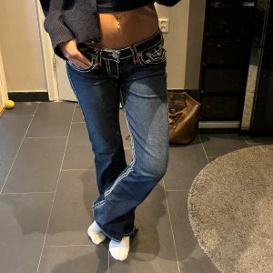 True Religion jeans - Säljer ett par blå True Religion jeans med vita och röda kontrastsömmar, låg midja och bootcut-ben.  Har slitningar längst ner på benet (se bild), därav priset. Skriv för frågor eller mer bilder💕