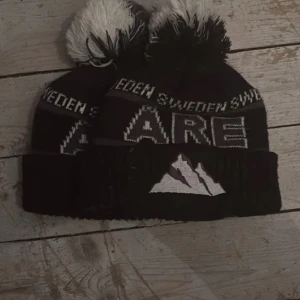 Svart Åre mössa med tofs - Säljer en svart stickad Åre-mössa med vit och grå text 'Sweden' och 'Åre' samt broderat bergmotiv framtill. Mössan har en stor svartvit tofs på toppen och uppvikt kant. Perfekt för kalla dagar i fjällen eller stan.